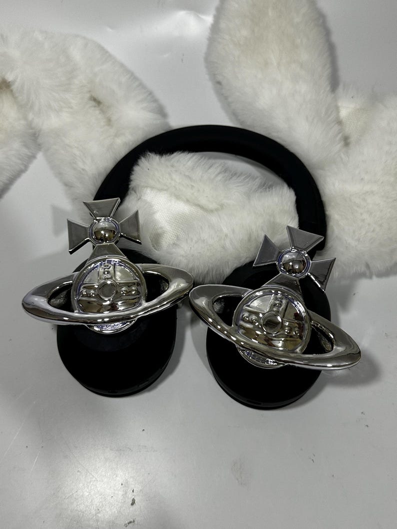 May include: A pair of black headband earmuffs with white faux fur ear covers. Each earmuff features a silver orb with a cross and a ring around it.