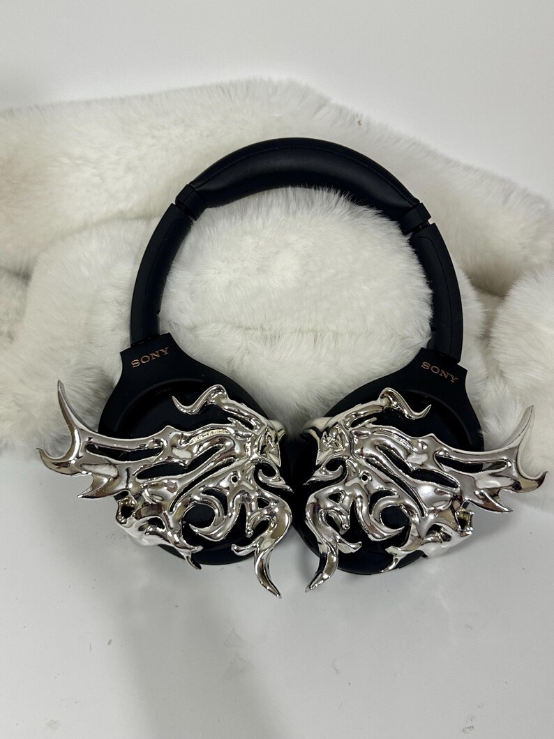 May include: Black Sony headphones with silver, ornate, wing-like embellishments. The headphones rest on a white, fluffy surface. The earcups feature the Sony logo.