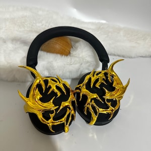 May include: Black headphones with gold-colored, intricate, decorative earcups. The earcups have a design that resembles a stylized, abstract flame.