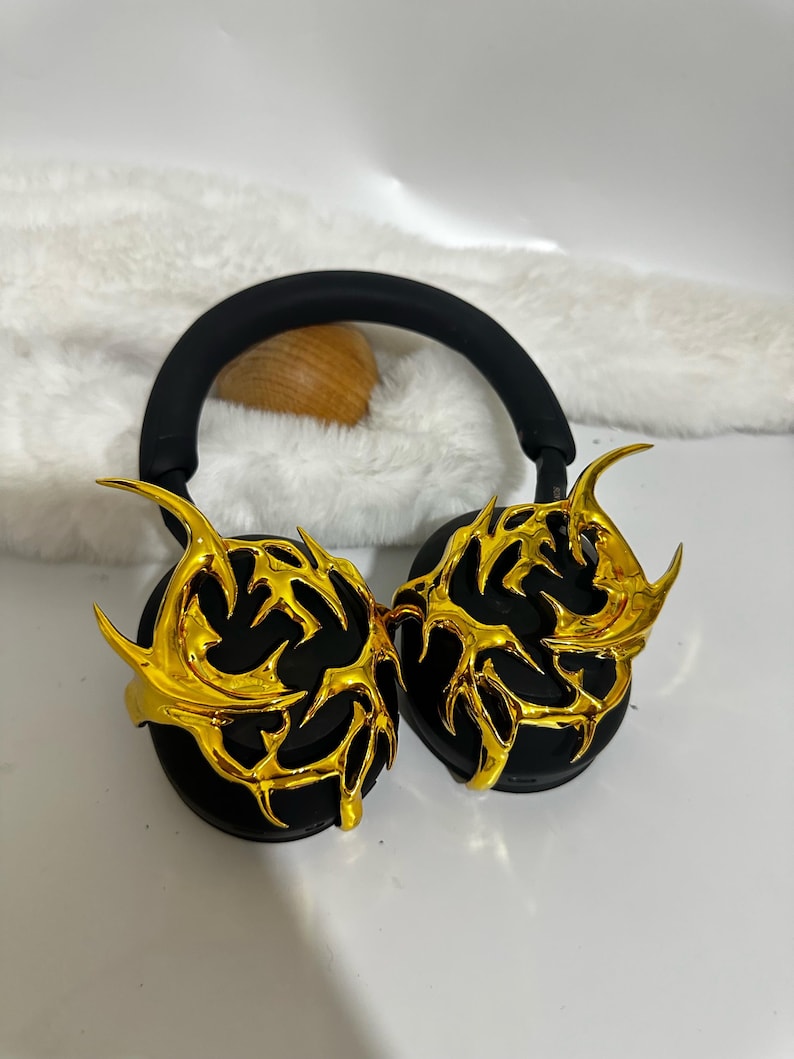 May include: Black headphones with gold-colored, intricate, decorative earcups. The earcups have a design that resembles a stylized, abstract flame.