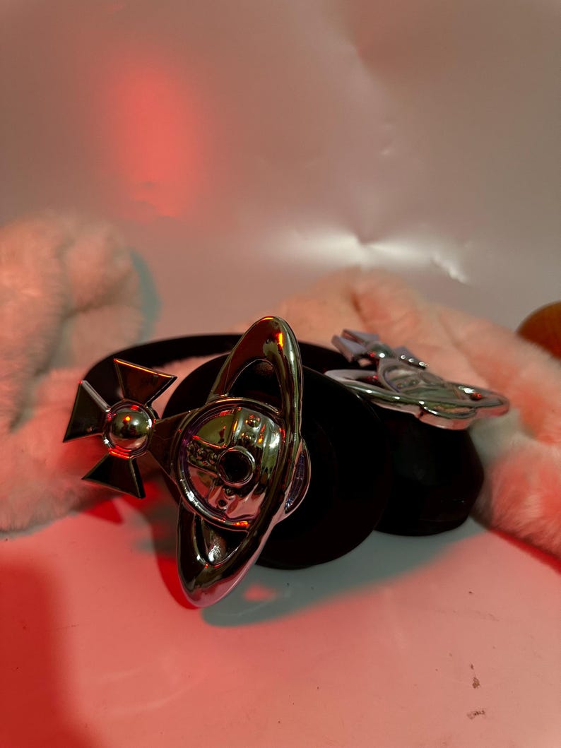 May include: A silver orb pendant with a cross and a ring design. The pendant is attached to a black leather strap.