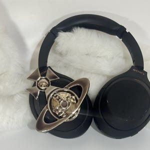 May include: Black Sony headphones with a silver decorative emblem. The headphones have a padded headband and earcups. The emblem is a silver-colored design with a planet-like shape.
