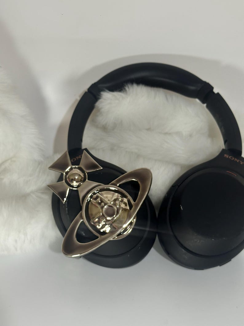 May include: Black Sony headphones with a silver decorative accent featuring a planetary design. The headphones are resting on a white, fluffy surface. The earcups are black and the headband is also black.