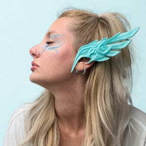 May include: A turquoise wing-shaped ear cuff is worn by a person in a side profile. The ear cuff has a detailed, feathered design. The person has blonde hair and light blue face paint. The background is a solid light blue.