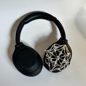 May include: Black Sony headphones with a silver spiderweb design on one ear cup. The headphones are on a white surface. The Sony logo is visible on the ear cups.
