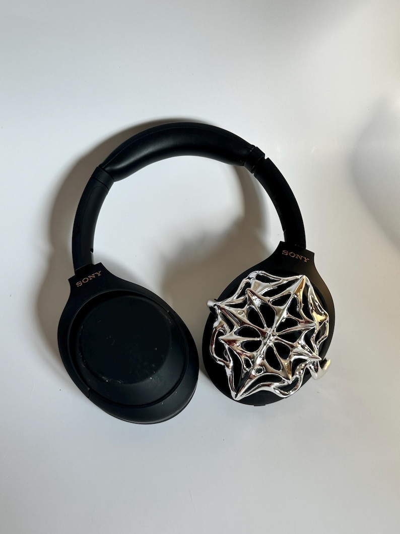 May include: Black Sony headphones with a silver spiderweb design on one ear cup. The headphones are on a white surface. The Sony logo is visible on the ear cups.