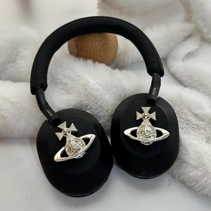 Puede incluir: Auriculares negros de diadema con detalles plateados. Los auriculares tienen una diadema y almohadillas acolchadas. Los auriculares presentan un dise&ntilde;o plateado de orbe y cruz. Los auriculares est&aacute;n sobre una superficie blanca y esponjosa.