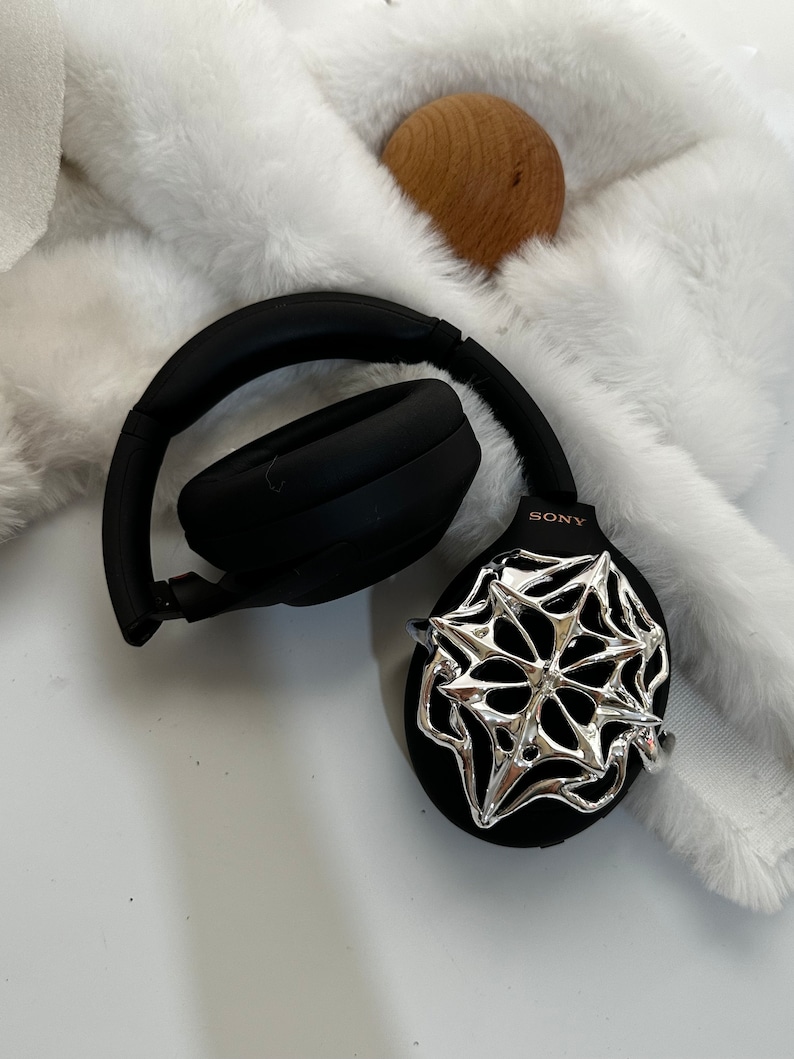 May include: Black over-ear headphones with a silver decorative accent. The headphones are resting on a white, fluffy surface. The brand name "SONY" is visible on the side of the headphones. A wooden sphere is also in the image.