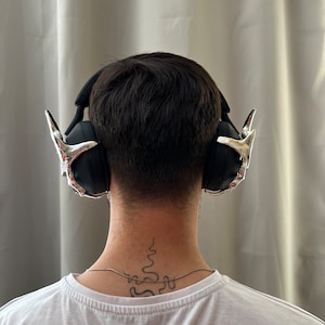 May include: A person wearing black headphones with silver accents. The headphones have a unique design with pointed earcups. The person has a tattoo on the back of their neck.