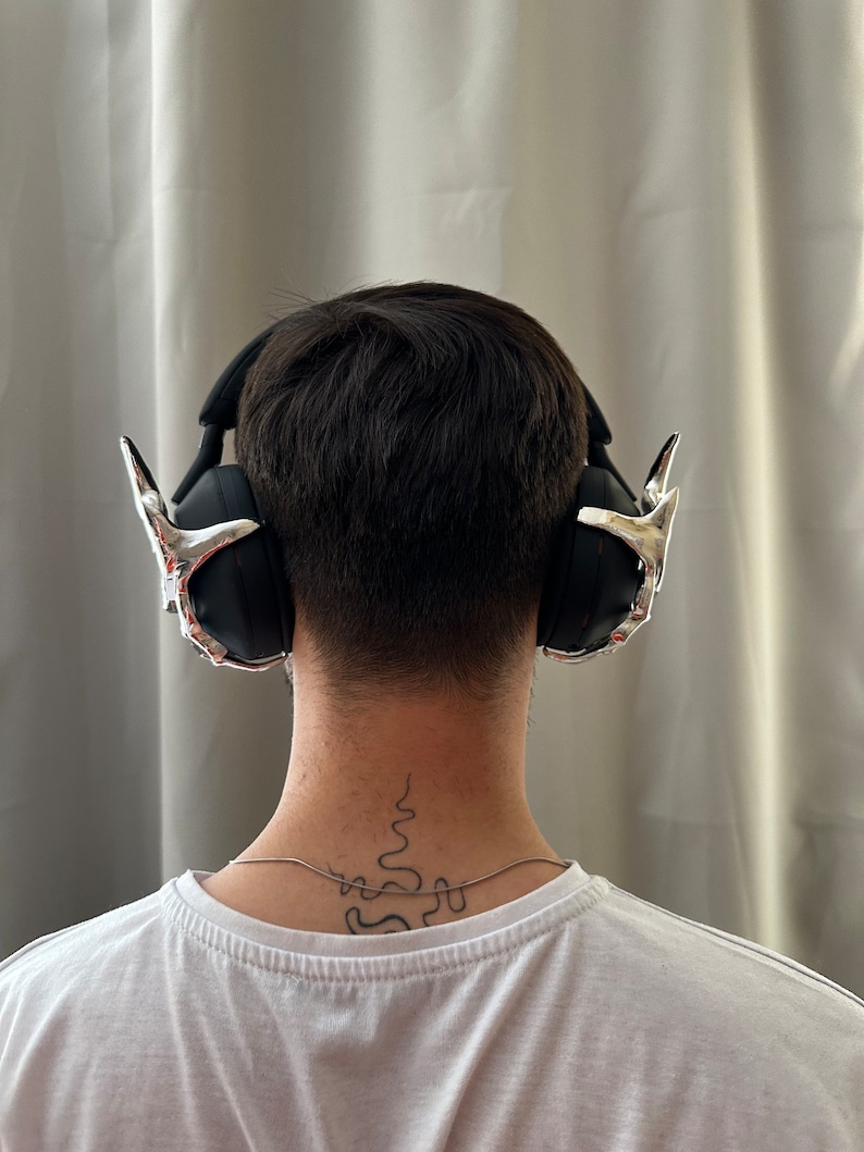 May include: A person wearing black headphones with silver accents. The headphones have a unique design with pointed earcups. The person has a tattoo on the back of their neck.