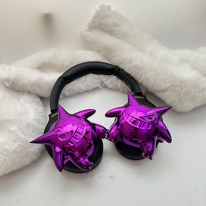 May include: Black Sony headphones with metallic purple Gengar figures attached to the earcups. The headphones have a black headband and earcups. The Gengar figures are a shiny, vibrant purple color and have a cartoonish design.
