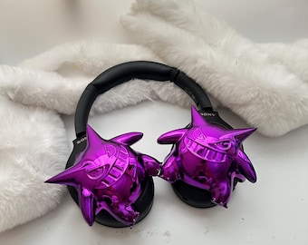 Può includere: Cuffie Sony nere con figure Gengar viola metallizzato attaccate ai padiglioni auricolari. Le cuffie hanno una fascia e padiglioni neri. Le figure di Gengar sono di un colore viola brillante e vibrante e hanno un design a cartoni animati.