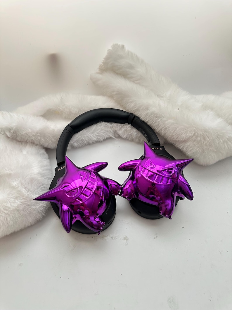 May include: Black Sony headphones with metallic purple Gengar figures attached to the earcups. The headphones have a black headband and earcups. The Gengar figures are a shiny, vibrant purple color and have a cartoonish design.