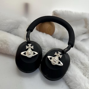 Puede incluir: Auriculares negros de diadema con adornos plateados de orbe y cruz en cada auricular. Los auriculares tienen una diadema y almohadillas negras acolchadas. El fondo es una textura suave y blanca.