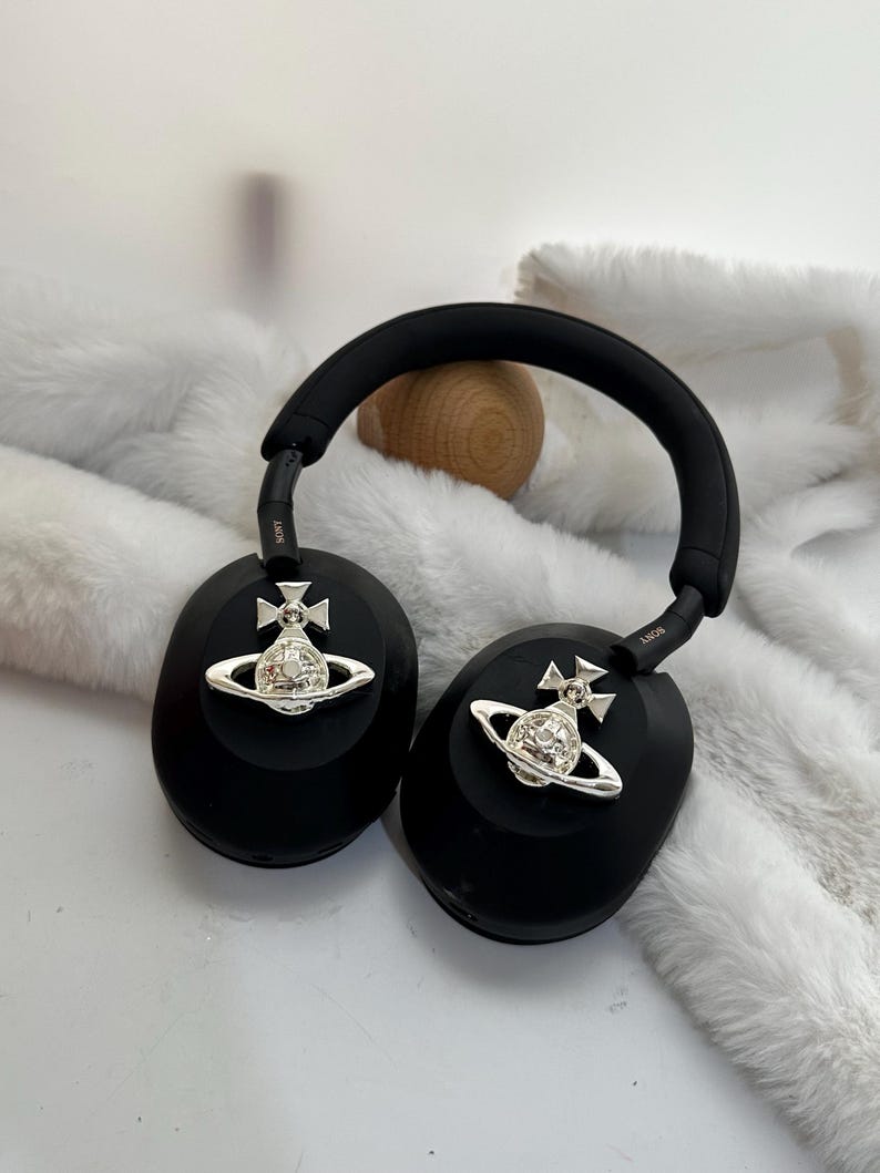 Puede incluir: Auriculares negros de diadema con adornos plateados de orbe y cruz en cada auricular. Los auriculares tienen una diadema y almohadillas negras acolchadas. El fondo es una textura suave y blanca.