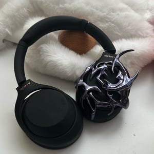 Może przedstawiać: Czarne słuchawki nauszne z ozdobnym, metalicznym, abstrakcyjnym wzorem na jednym nauszniku. Słuchawki mają napis "SONY" nadrukowany na obu nausznikach.