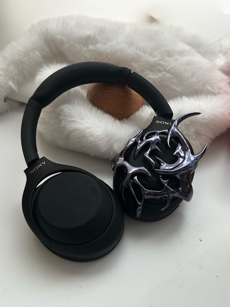 Może przedstawiać: Czarne słuchawki nauszne z ozdobnym, metalicznym, abstrakcyjnym wzorem na jednym nauszniku. Słuchawki mają napis "SONY" nadrukowany na obu nausznikach.