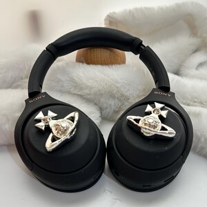 Puede incluir: Auriculares Sony negros con detalles plateados. Los auriculares presentan un dise&ntilde;o plateado de orbe y cruz. Los auriculares descansan sobre una superficie blanca y esponjosa.