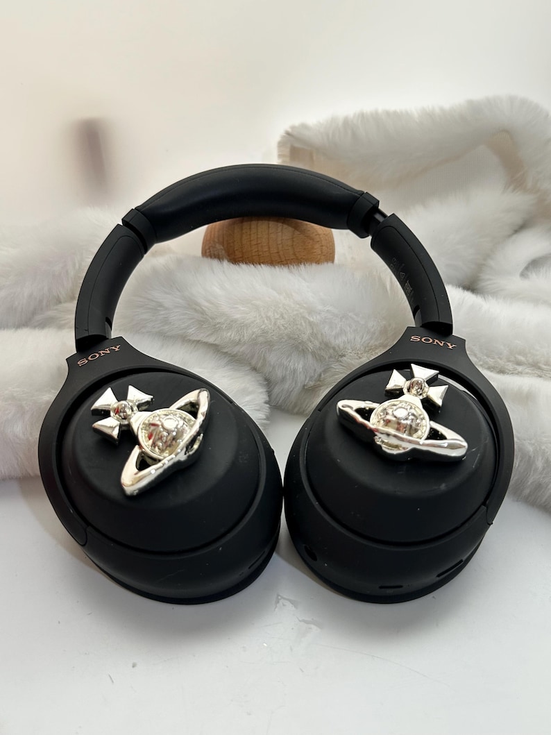Puede incluir: Auriculares Sony negros con detalles plateados. Los auriculares presentan un dise&ntilde;o plateado de orbe y cruz. Los auriculares descansan sobre una superficie blanca y esponjosa.