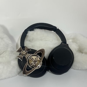 May include: Black Sony headphones with a silver decorative emblem featuring a cross and a planet design. The headphones rest on a white, fluffy surface, with the Sony logo visible on the ear cup.