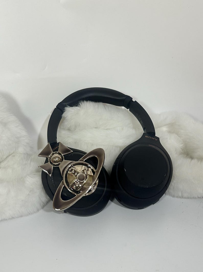 May include: Black Sony headphones with a silver decorative emblem featuring a cross and a planet design. The headphones rest on a white, fluffy surface, with the Sony logo visible on the ear cup.