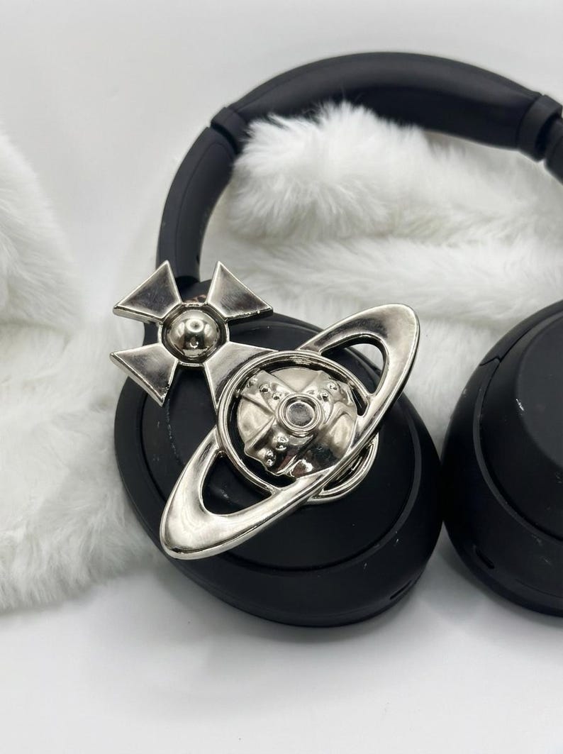 May include: Black headphones with a silver emblem featuring a cross and a ring design. The headphones rest on a white, fluffy surface. The emblem is a prominent detail, adding a decorative element to the audio accessory.