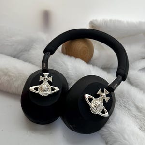 Puede incluir: Auriculares negros de diadema con adornos plateados. Los auriculares tienen una diadema y almohadillas acolchadas. Cada auricular tiene un emblema plateado. Los auriculares est&aacute;n sobre una superficie blanca y peluda.