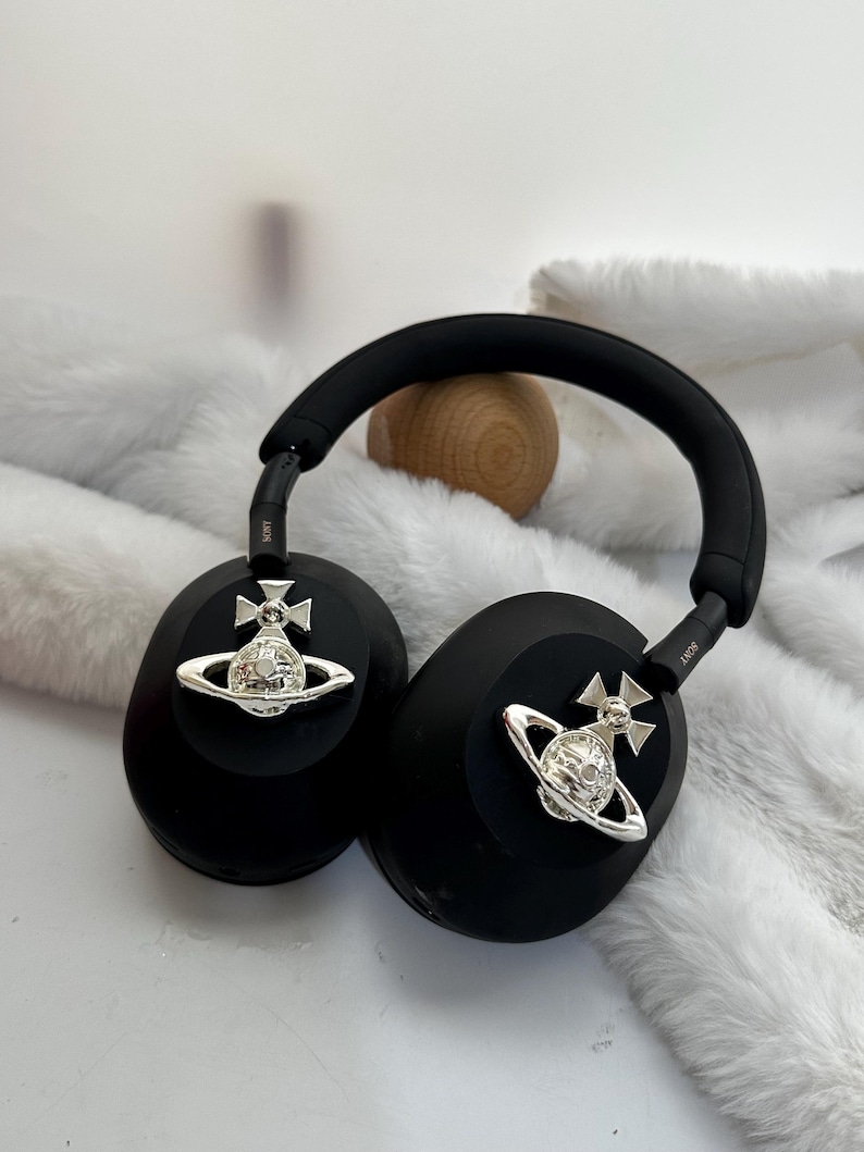 Puede incluir: Auriculares negros de diadema con adornos plateados. Los auriculares tienen una diadema y almohadillas acolchadas. Cada auricular tiene un emblema plateado. Los auriculares est&aacute;n sobre una superficie blanca y peluda.