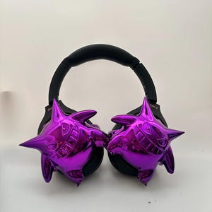 May include: Black over-ear headphones with shiny purple cartoon character designs on each ear cup. The headphones have a black headband and ear pads. The character design has a grinning face with sharp teeth and pointed ears.