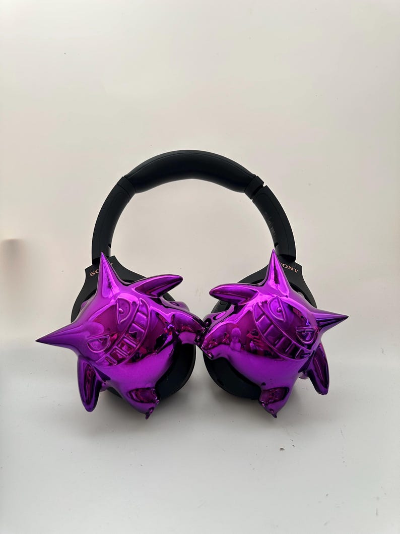May include: Black over-ear headphones with shiny purple cartoon character designs on each ear cup. The headphones have a black headband and ear pads. The character design has a grinning face with sharp teeth and pointed ears.