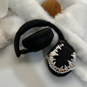 May include: Black wireless headphones with a silver, sharp-toothed design on one earcup. The headphones are on a white fluffy surface. The text "SONY" is visible on the earcup.