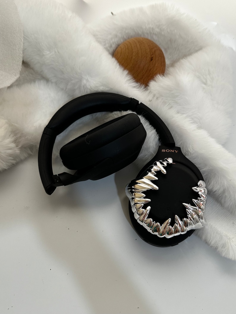 May include: Black wireless headphones with a silver, sharp-toothed design on one earcup. The headphones are on a white fluffy surface. The text "SONY" is visible on the earcup.