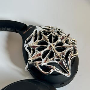 May include: Black Sony headphones with a silver, star-shaped decorative accent. The headphones are on a white surface. The word "SONY" is visible on the side of the headphones.