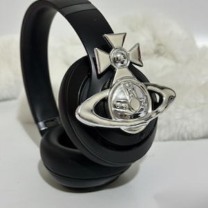 Op de afbeelding: Zwarte over-ear hoofdtelefoon met een zilveren embleem met een kruis en een planeetontwerp. De hoofdtelefoon heeft een gewatteerde hoofdband en oorschelpen. Het embleem is een opvallend kenmerk en voegt een vleugje luxe toe.