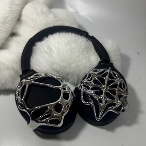 May include: Black over-ear headphones with silver decorative accents. The headphones have a black headband and earcups, with intricate silver designs on the earcups. The background features a fluffy white material.