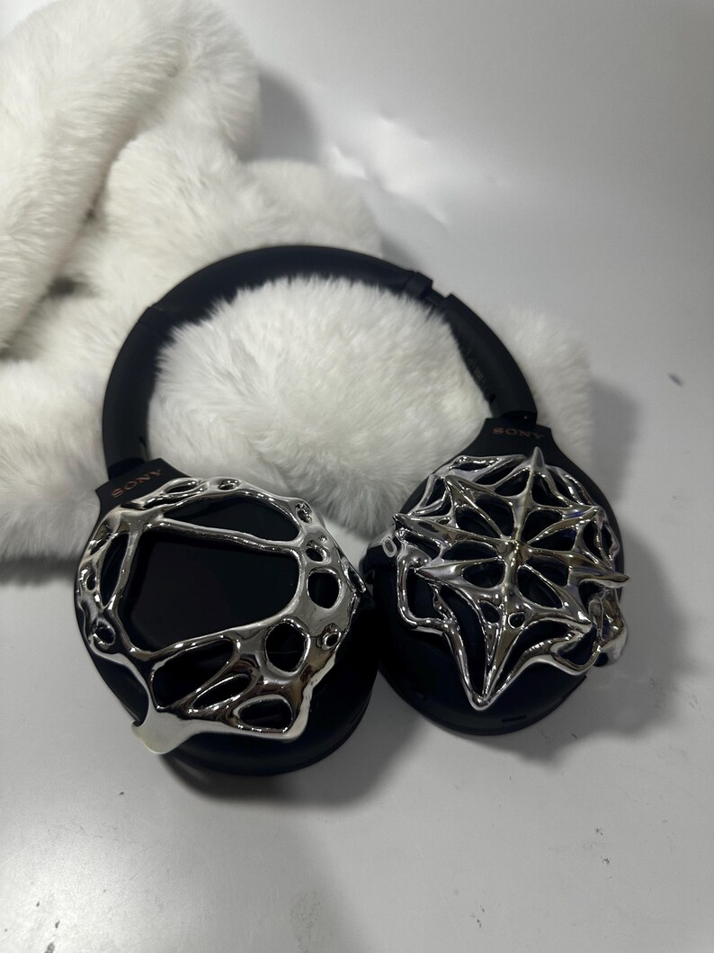May include: Black over-ear headphones with silver decorative accents. The headphones have a black headband and earcups, with intricate silver designs on the earcups. The background features a fluffy white material.