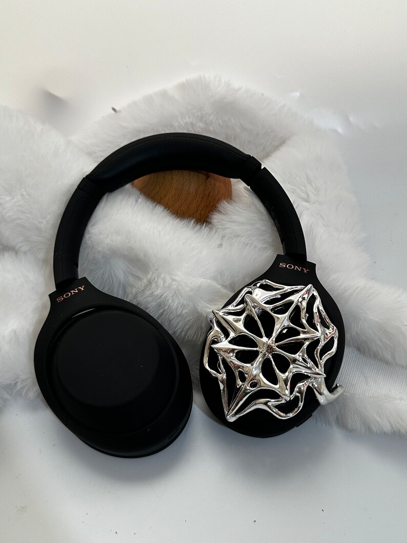 May include: Black Sony headphones with a silver spiderweb design on one ear cup. The headphones have a black headband and earcups, with the Sony logo in a rose gold color. The headphones are resting on a white, fluffy surface.