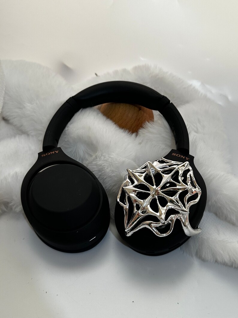 May include: Black Sony headphones with a silver, abstract design on one ear cup. The headphones are resting on a white, fluffy surface. The Sony logo is visible on the ear cups.