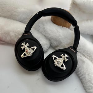 Puede incluir: Auriculares negros de diadema con detalles plateados. Cada auricular tiene un emblema plateado. La marca "SONY" est&aacute; impresa en los auriculares. Los auriculares est&aacute;n sobre una superficie blanca.