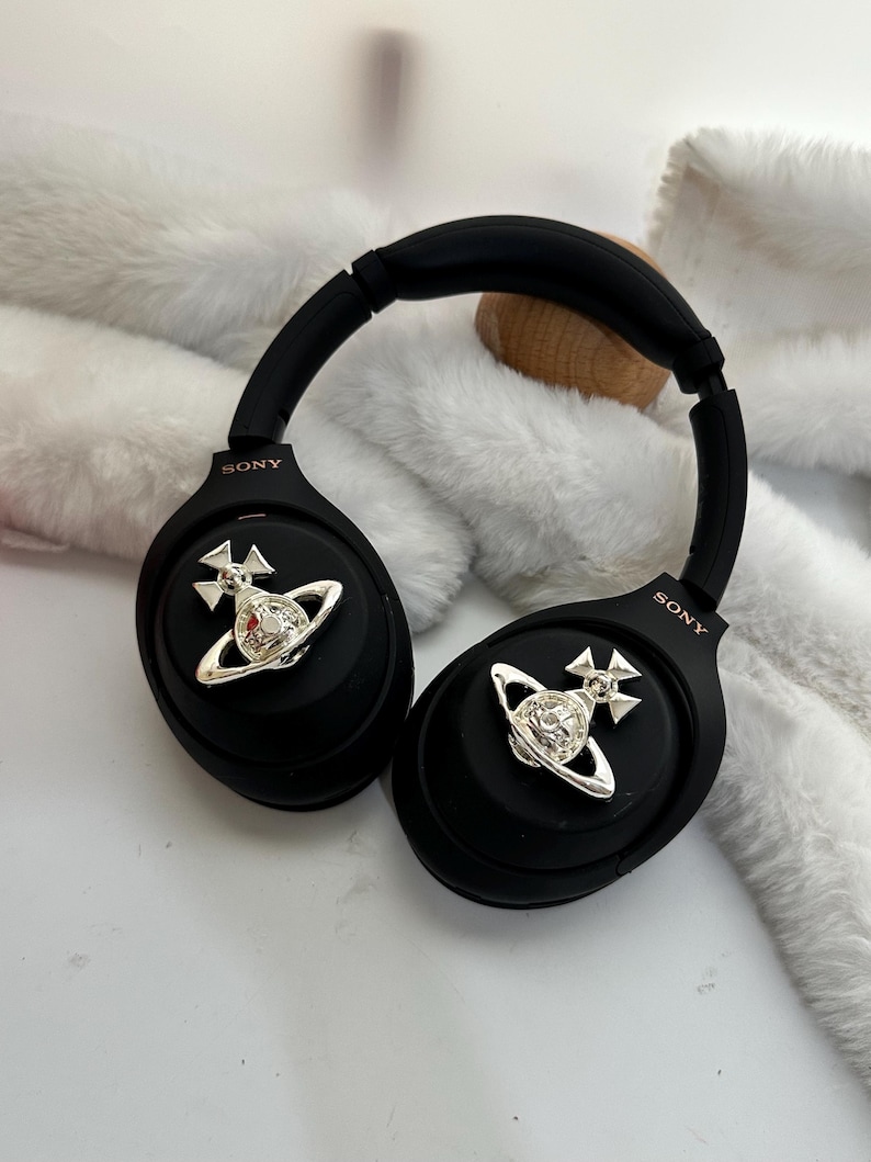 Puede incluir: Auriculares negros de diadema con detalles plateados. Cada auricular tiene un emblema plateado. La marca "SONY" est&aacute; impresa en los auriculares. Los auriculares est&aacute;n sobre una superficie blanca.