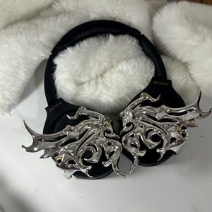 May include: Black Sony headphones with silver decorative accents. The headphones have a black headband and earcups. The silver accents feature an intricate, stylized design. The background includes white, fluffy material.