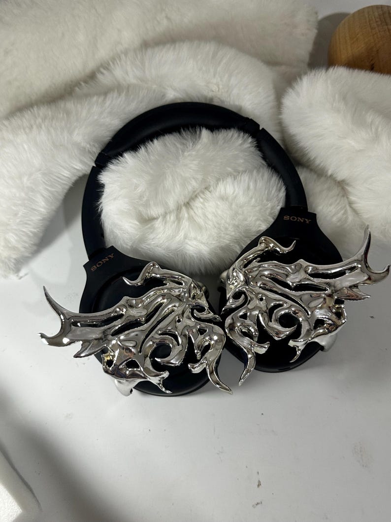 May include: Black Sony headphones with silver decorative accents. The headphones have a black headband and earcups. The silver accents feature an intricate, stylized design. The background includes white, fluffy material.
