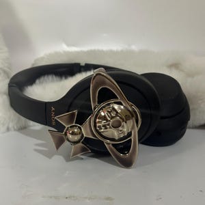 May include: Black Sony headphones with a silver decorative emblem. The emblem features a silver orb and ring design. The headphones are resting on a white, fluffy surface.