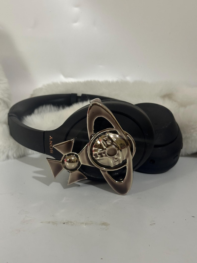 May include: Black Sony headphones with a silver decorative emblem. The emblem features a silver orb and ring design. The headphones are resting on a white, fluffy surface.