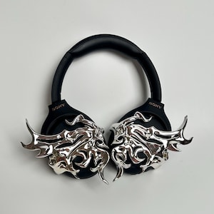 May include: Black Sony headphones with silver decorative accents. The accents feature an ornate, stylized design. The headphones are set against a white background.