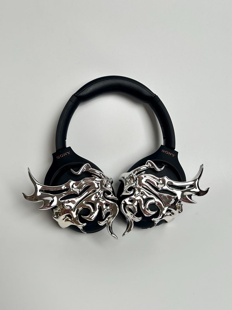 May include: Black Sony headphones with silver decorative accents. The accents feature an ornate, stylized design. The headphones are set against a white background.