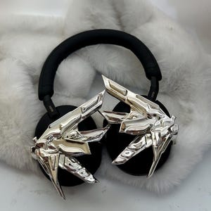 Puede incluir: Auriculares negros con detalles plateados en forma de manos estilizadas. Los auriculares tienen una diadema y auriculares negros. El fondo es un material blanco y esponjoso.