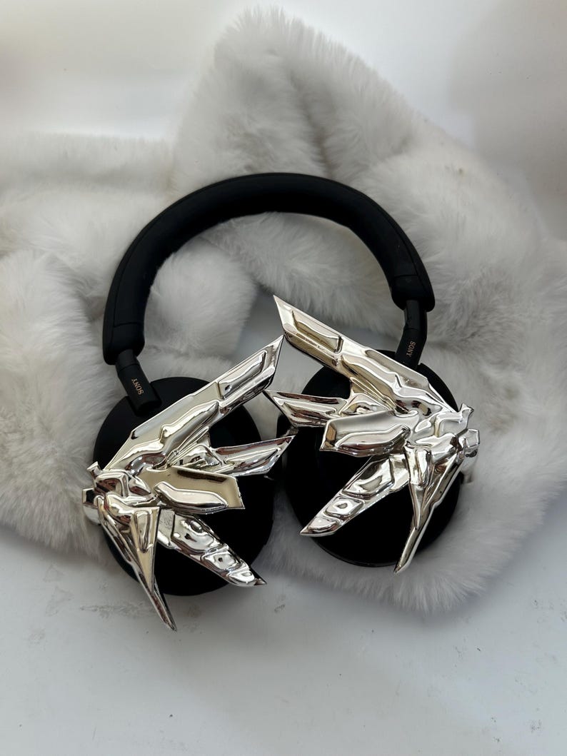 Puede incluir: Auriculares negros con detalles plateados en forma de manos estilizadas. Los auriculares tienen una diadema y auriculares negros. El fondo es un material blanco y esponjoso.