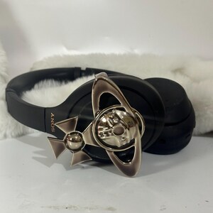 May include: Black Sony headphones with a silver-toned decorative emblem. The emblem features a stylized design with a central orb and a cross-like shape. The headphones are resting on a white, fluffy surface.