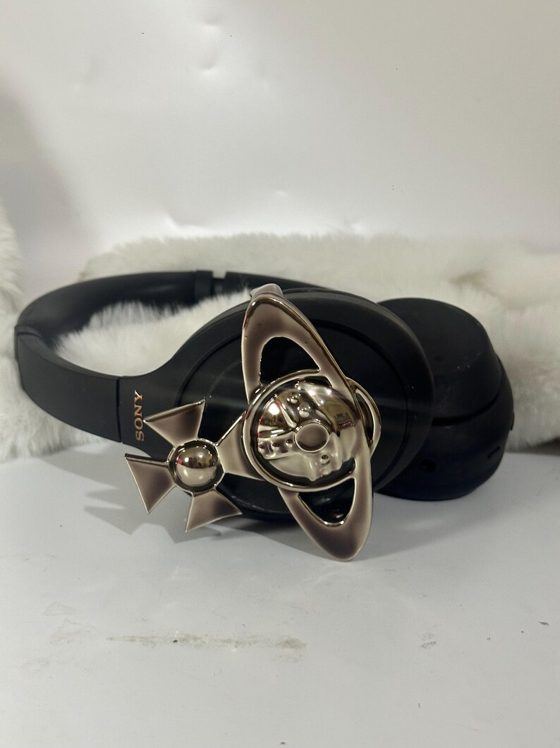 May include: Black Sony headphones with a silver-toned decorative emblem. The emblem features a stylized design with a central orb and a cross-like shape. The headphones are resting on a white, fluffy surface.